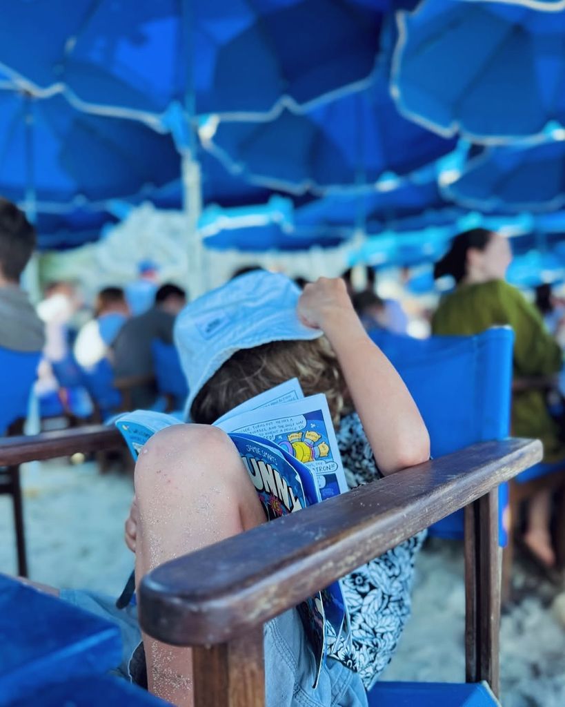 Cat Deeley's son wearing blue hat and matching outfit while reading his comic book on holiday