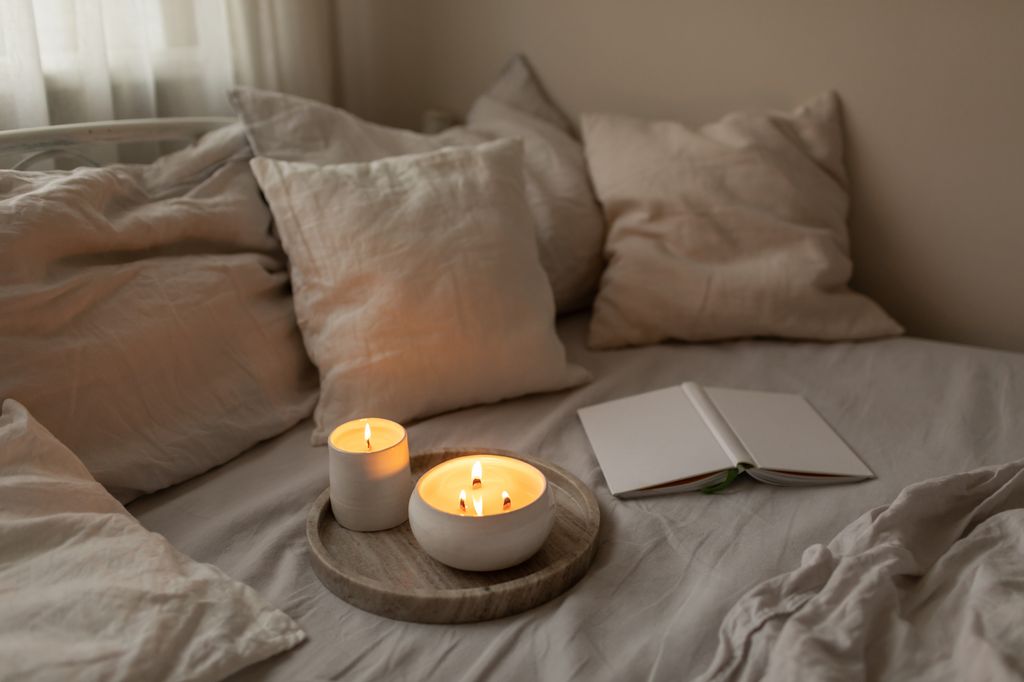 Organic soya wax candles with wooden candlewick in white ceramic bowls on linen bedding set with book. Cosy and soft winter background. Hygge and aromatherapy concept.