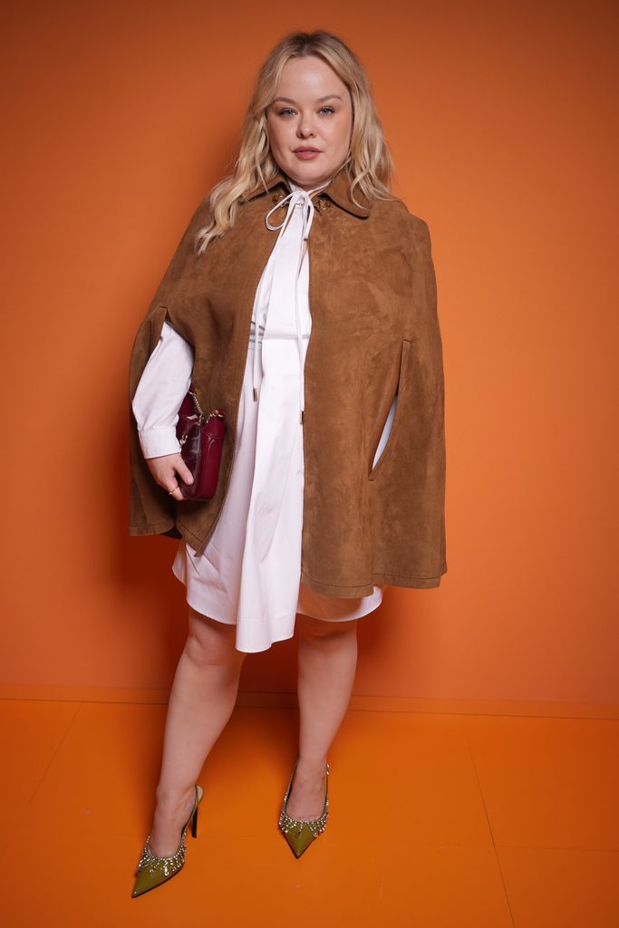 Nicola Coughlan wears an elegant, oversized brown suede cape over a white mini-dress, adding a sophisticated touch to her look. She holds a burgundy handbag in one hand, and her outfit is completed with pointed green heels adorned with crystal embellishments. She exudes confidence with soft makeup and wavy blonde hair.