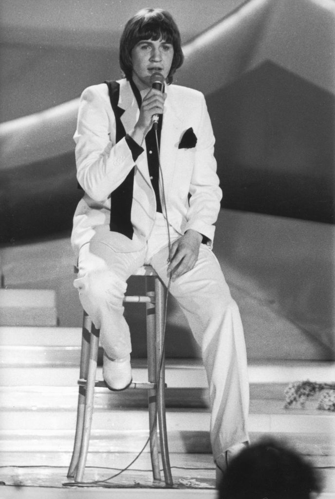 Johnny Logan wears a white tuxedo with a black shirt and pocket square while performing seated on a stool at Eurovision 1980.