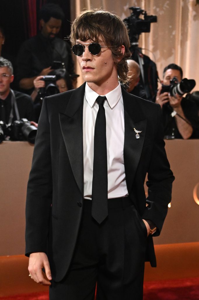 Connor Storrie at the Golden Globes red carpet wearing a black suit, white shirt and black tie, teamed with retro sunglasses