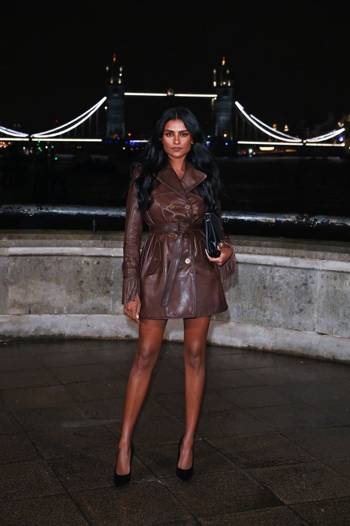Simone Ashley porte un trench-coat en cuir marron avec un sac à main et des talons noirs