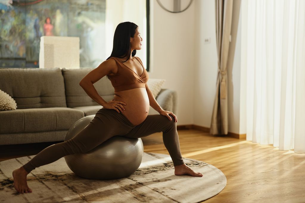Smiling Asian expecting woman doing relaxation exercises on fitness ball in the living room.