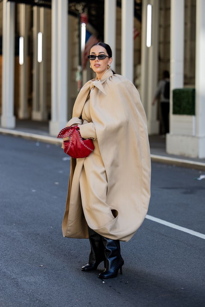 Olga Sokolenko wears red bag, beige cape / trench coat, black boots, sunglasses outside during New York Fashion Week on September 10, 2024 in New York City.