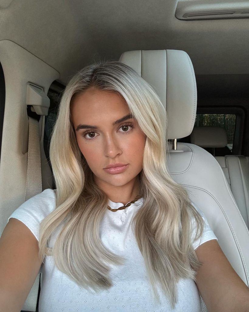 Molly-Mae's sits in a car and poses for a selfie. Her hair is parted in the middle, and a very light, glossy and expensive looking shade of buttermilk blonde