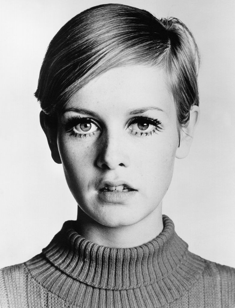 Neste retrato atemporal em preto e branco, a expressão de olhos arregalados e o corte pixie característico de Twiggy tomam o centro do palco. A gola alta minimalista complementa suas características marcantes, acentuando os cílios ousados ​​que se tornaram seu visual definidor. Um rosto que sintetizou o movimento mod dos anos 60, esta imagem a imortaliza como um verdadeiro ícone da moda.