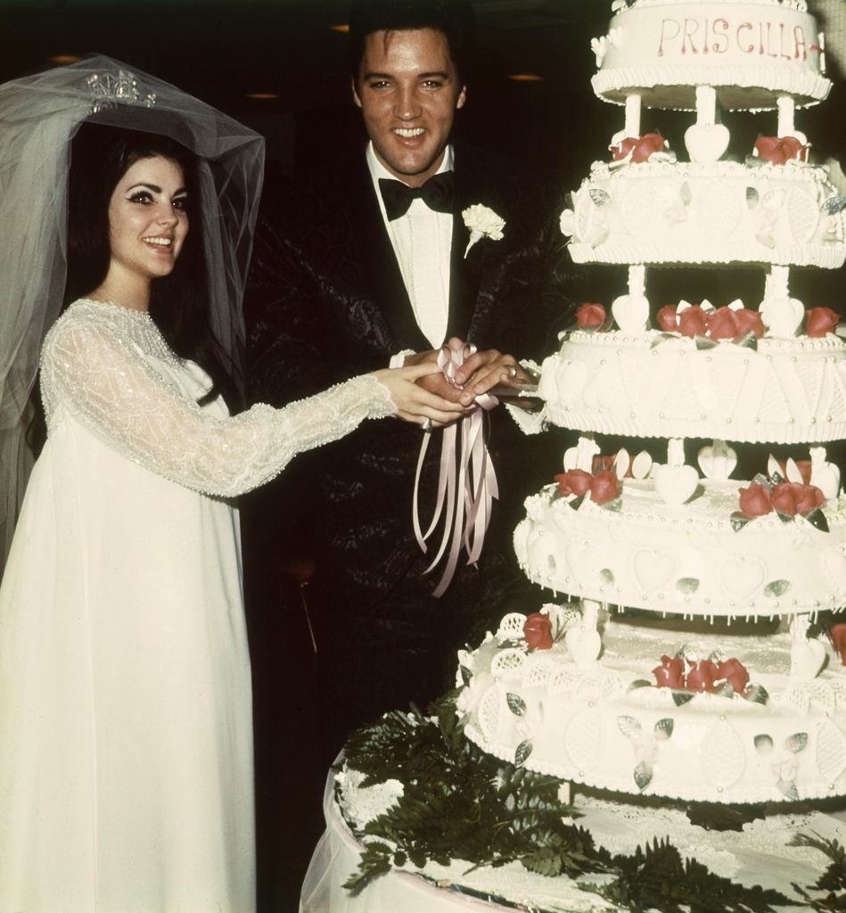 Priscilla Presley wearing a floaty wedding dress while cutting her wedding cake with Elvis