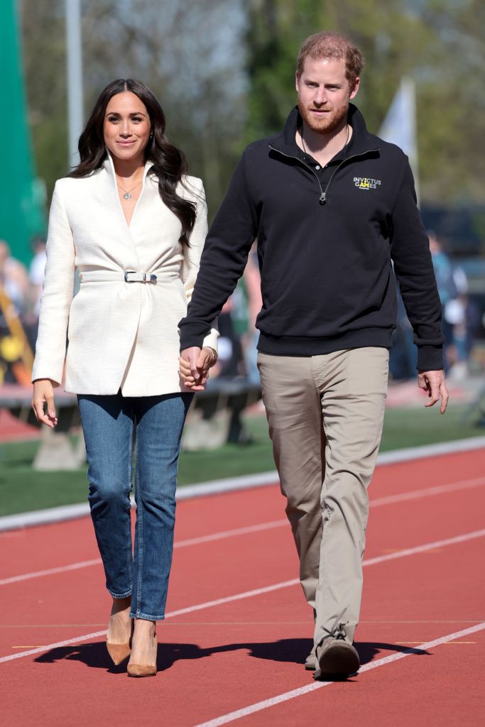 Meghan Markle in a white coat and jeans walking with Prince Harry in a black jacket and tan trousers