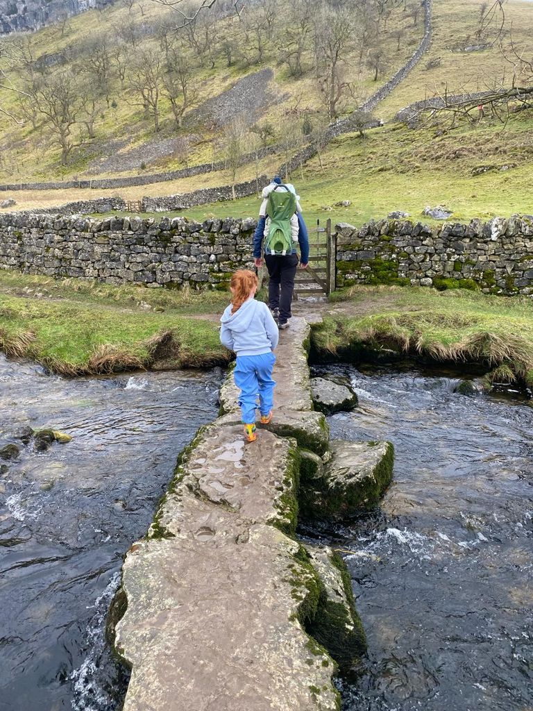 Man with baby on back and red-haired girl cross a stone bridge over water