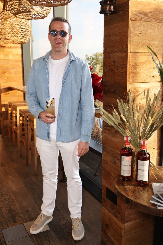 Harry allemand tient le classique dans une chemise en chambre pâle, un jean blanc, des baskets en daim gris et des lunettes de soleil rondes tout en profitant de cocktails lors d'un événement de whisky éclairé par le soleil organisé par Gentlemen's Journal et Star Hill Farm