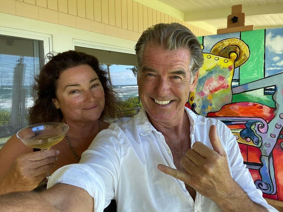 Pierce Brosnan smiling in a white shirt next to his wife drinking wine