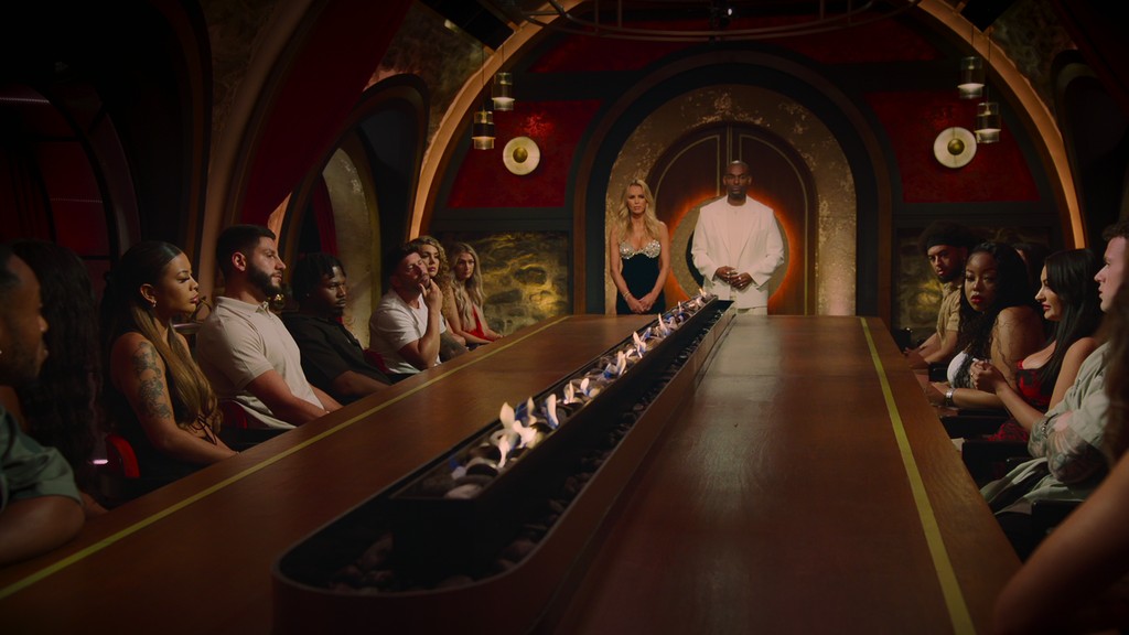 female and male tv hosts in front of table of contestants 