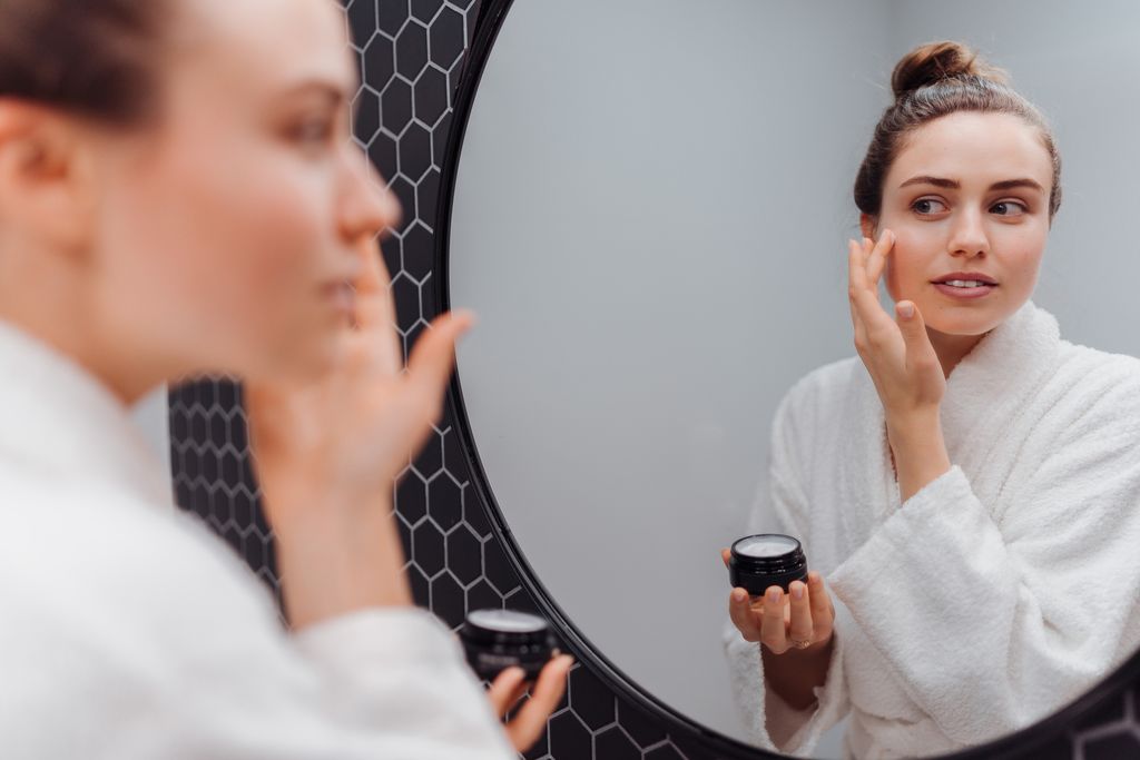 Young woman in bathrobe putting on cream in the mirror