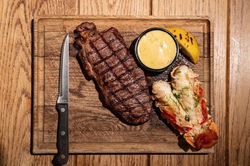 A grilled steak, lobster tail, and sauce on a wooden board with a steak knife at The Bountiful Cow, Holborn, London