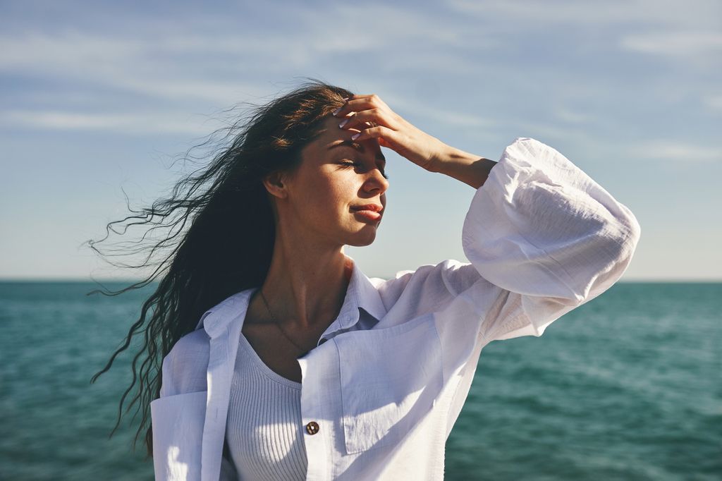 Une femme debout à côté de la mer avec sa main sur son front, l'air réfléchie. Ses cheveux soufflent dans le vent. Le ciel est clair et la mer est en arrière-plan.