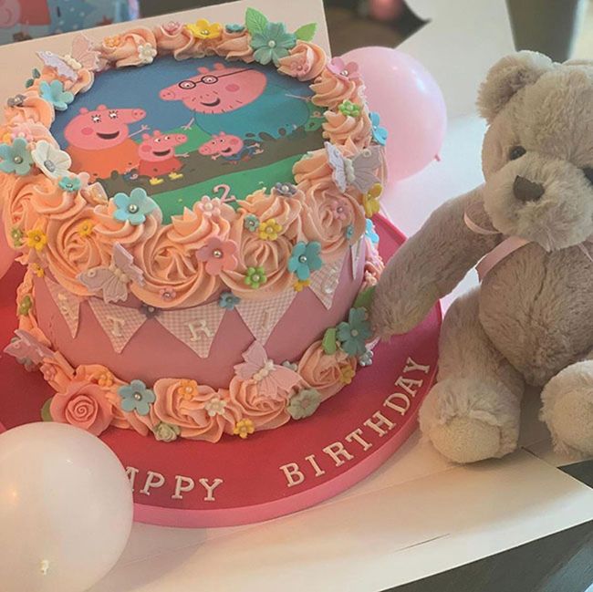 a pink cake decorated with pink swirls and a peppa pig cartoon picture is placed next to a light brown teddy bear