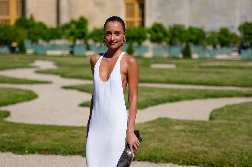 Alexandra Saint Mleux wears a white floor-length halterneck dress with a plunging neckline, deep side cutouts and a flared train hem, styled with gold sculptural hoop earrings and a black textured leather clutch bag. Hair is pulled back into a sleek bun, outside Jacquemus, during the Paris Fashion Week