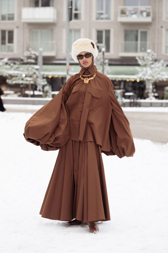Rawdah Mohamed wears brown long skirt and a wide puffy sleeve top, golden jewellery and beige fur hat outside the Forza Collective show during Copenhagen Fashion Week AW26