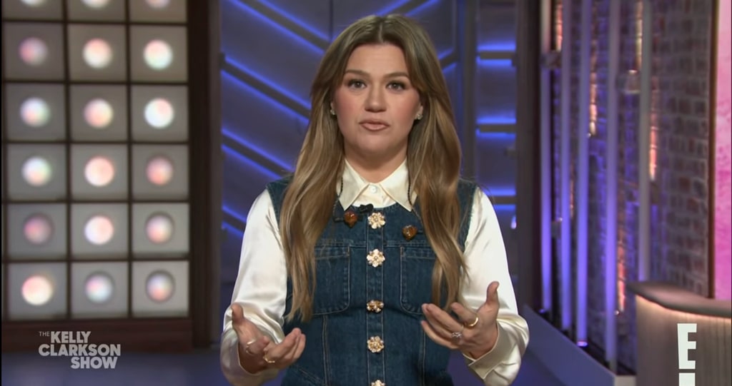 Kelly Clarkson wearing white shirt and denim dress while speaking to camera on The Kelly Clarkson Show