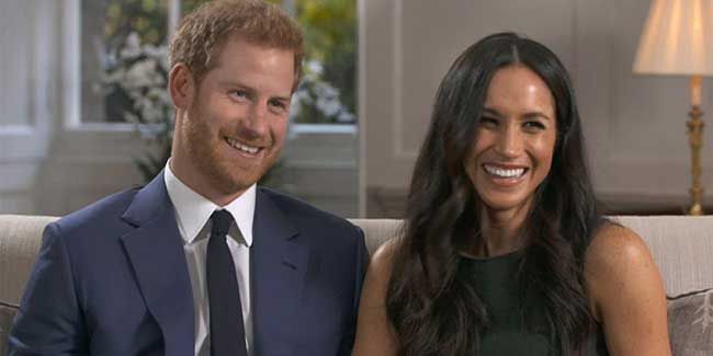 a mid shot of harry and meghan seated next to each other on a sofa smiling at someone positioned off camera