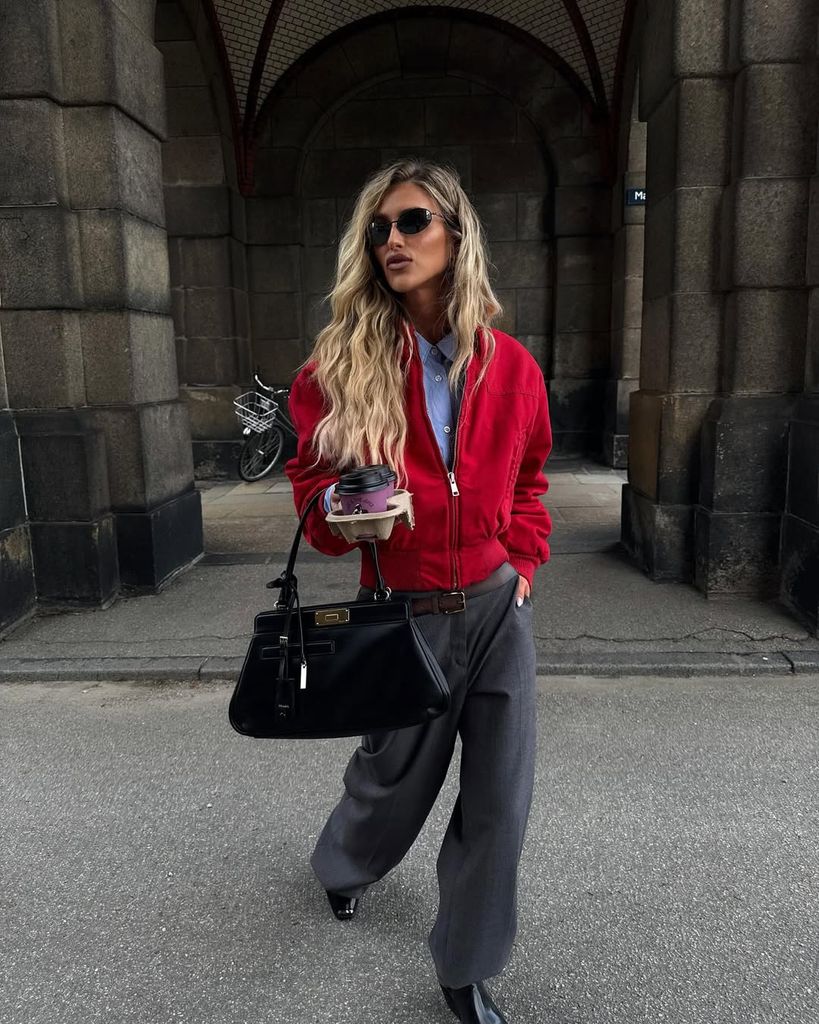 Danish content creator Emili Sindlev rocks a bold red bomber jacket, grey wide-leg trousers, and black accessories while clutching a takeaway coffee under arched stone architecture.