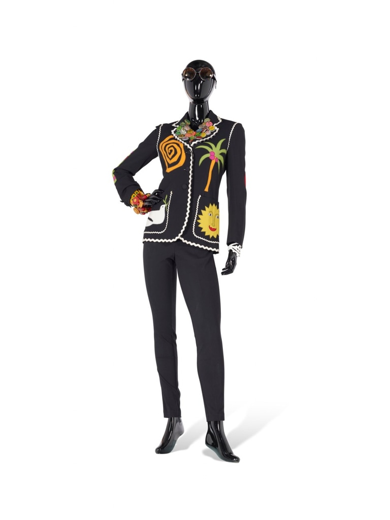  black jacket by Moschino Cheap and Chic (1995), adorned with colorful appliques of a palm tree, sun, and spiral. The jacket is paired with sleek black pants and accessorized with bold, chunky jewelry, including a colorful necklace. Oversized sunglasses complete the look. This piece belonged to the legendary Iris Apfel.