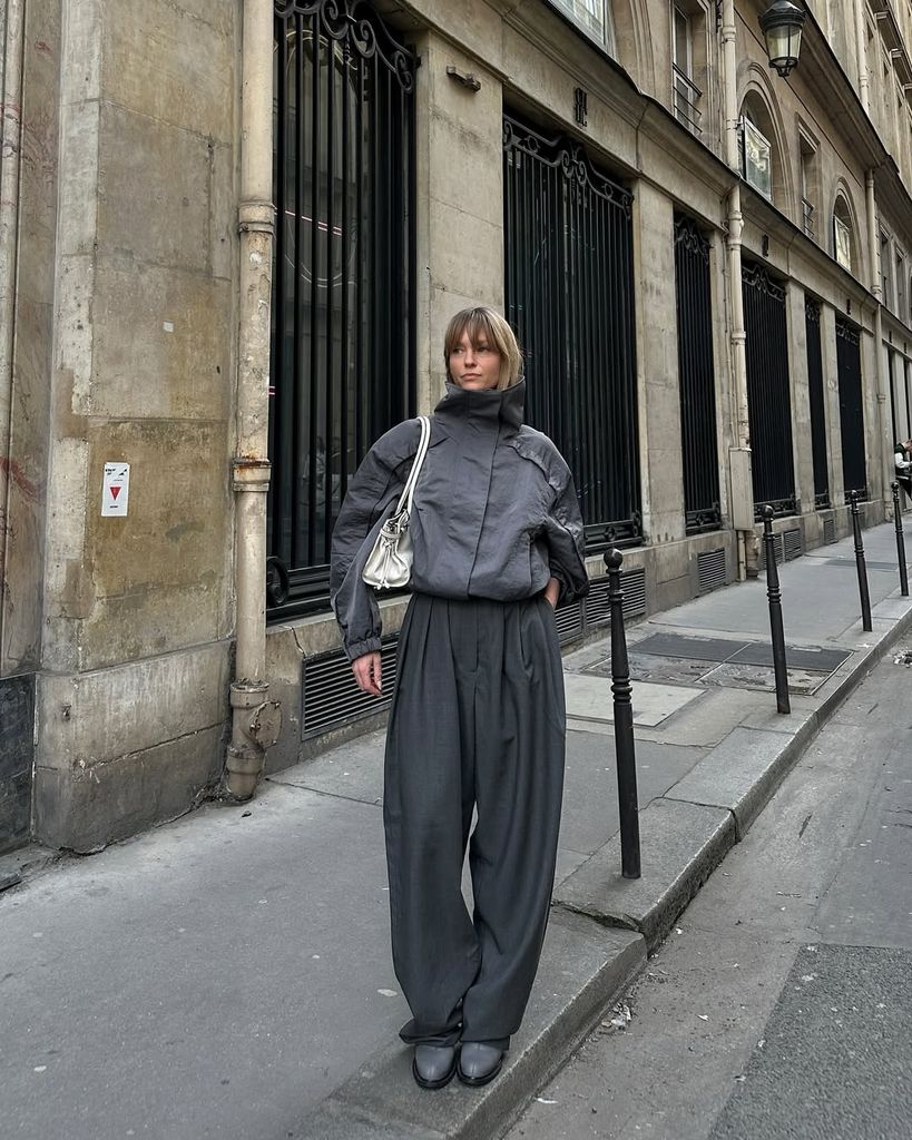 Danish fashion girl Jeanette Madsen stuns in oversized grey outerwear, pleated trousers and sleek boots while leaning against a historic Parisian building. Minimalist, moody, and undeniably cool.