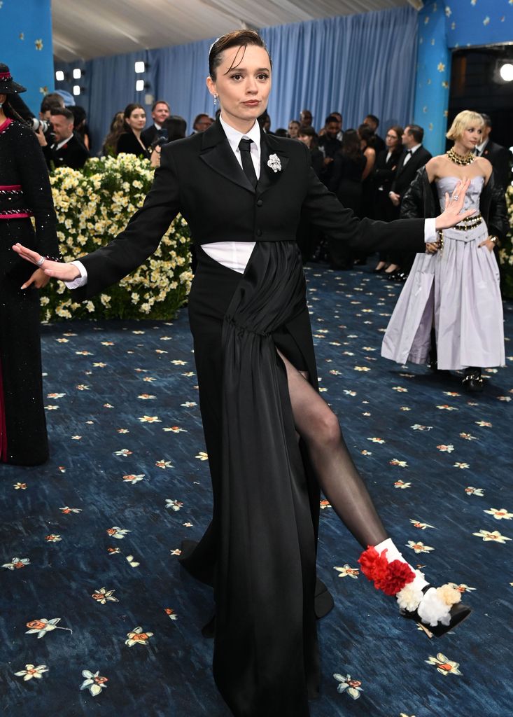 Aimee Lou Wood wears a cropped suit jacket, white shirt and tie, a long asymmetric skirt and heels embellished with white, cream and red flowers
