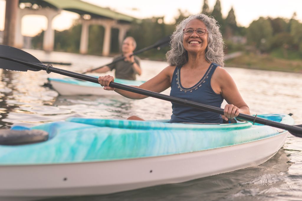 A mixed race senior couple is kayaking down a river together. The image's focus is on the elderly woman smiling in the foreground. She is Pacific Islander and has graying hair. Her husband is seen in a kayak in the background. In the distance behind the couple you can see the shoreline and a bridge. The happy couple are wearing casual clothing. It is warm outside.