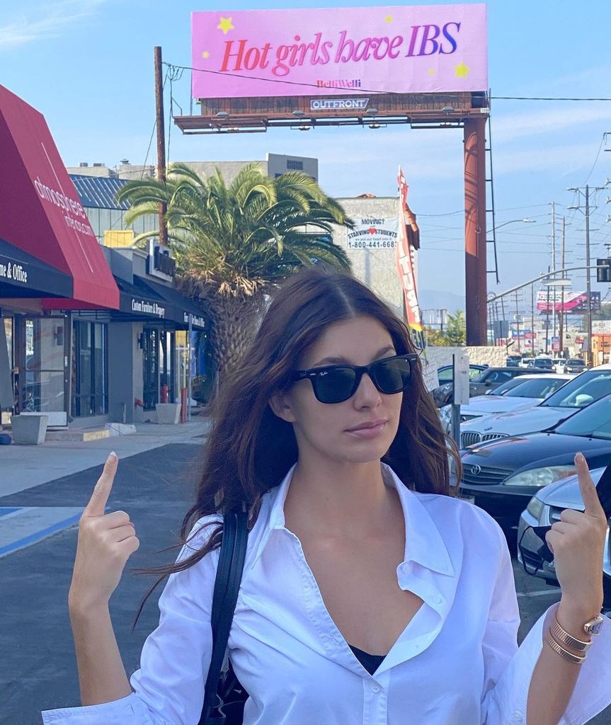 Daisy Jones & The Six star Camila Morrone posing beside a billboard that reads 'Hot Girls Have IBS'