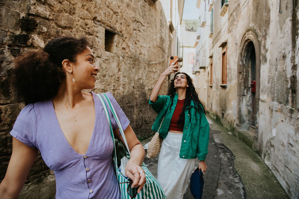 Two woman explore the back streets of a European city. The woman in the foreground glances over her shoulder and smiles at her friend, who uses her mobile phone to take a video of her surroundings.