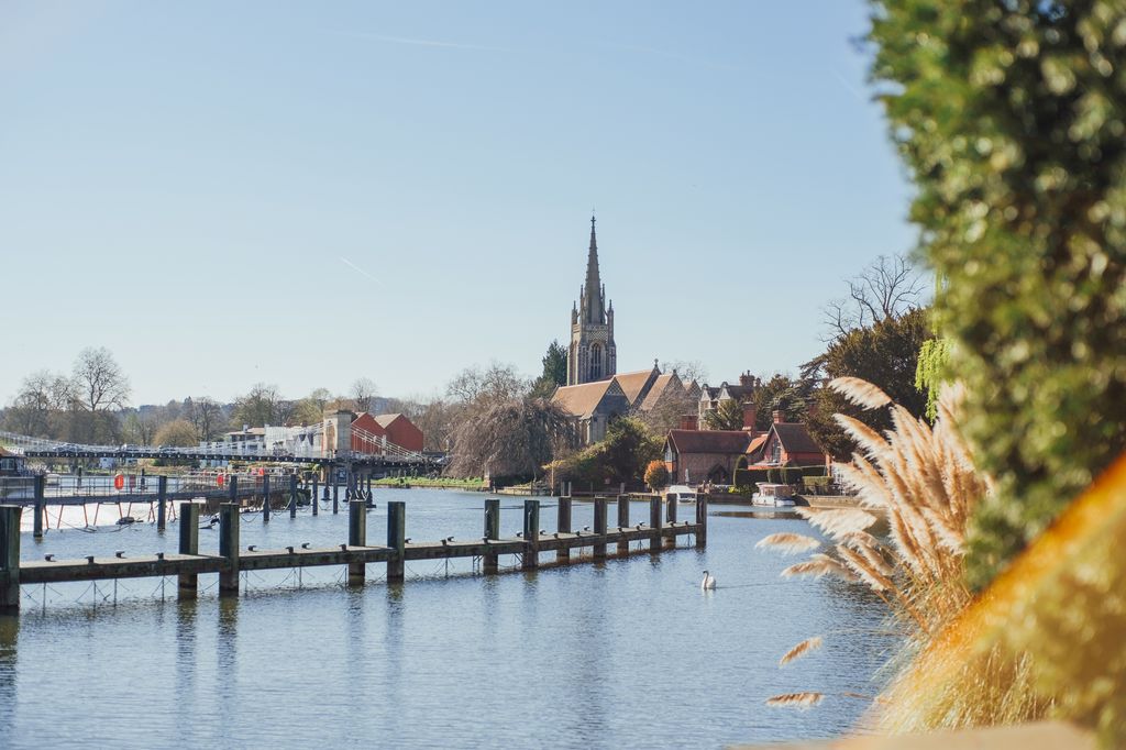 A scenic view of All Saints Church in Marlow, Buckinghamshire, England, from across the River Thames on a sunny day. The church's spire dominates the skyline.