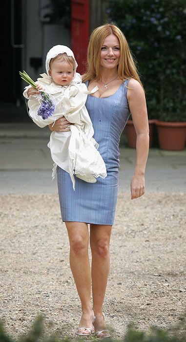 geri stands in a light blue dress holding a baby wearing white christening robes while clutching a bunch of bluebells