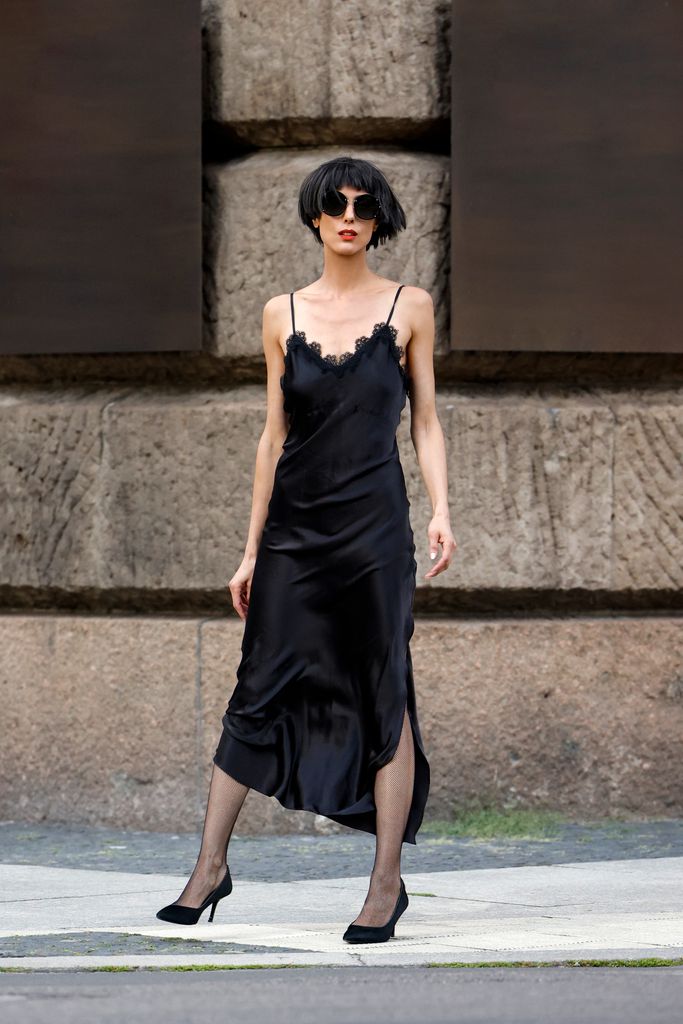 Model and artist Zoe Helali wearing the black "u2018Elisabeth"u2019 slip silk dress with shoulder straps, lace detail and a high one side slit by Sasha La Mer, black pumps by Dior, black round sunglasses by Dior and black fishnet tights by Wolford during a street style shooting on October 23, 2021