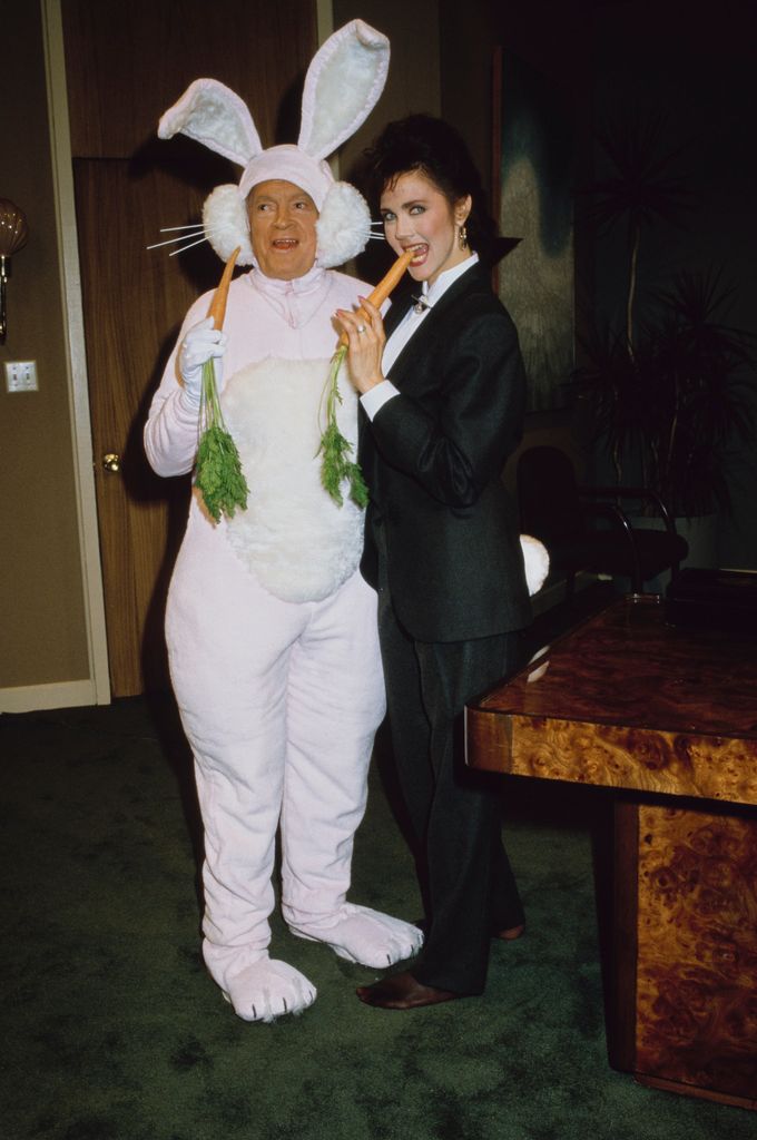 Bob Hope in a full bunny suit holds carrots beside Lynda Carter in a black pantsuit biting one.