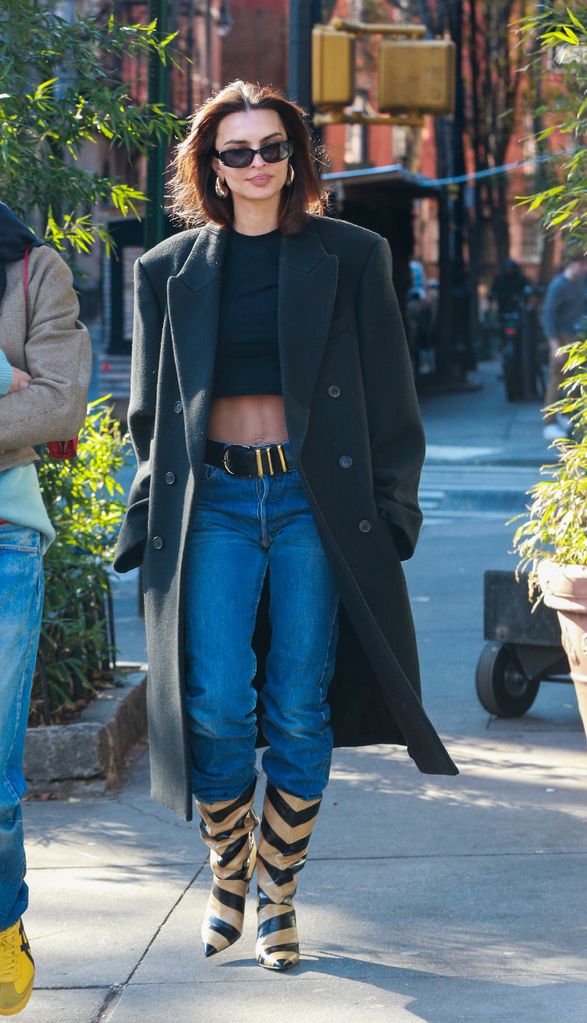 Emily Ratajkowski wears a pair of striped boots, jeans, a crop top and long over coat whilst in NYC