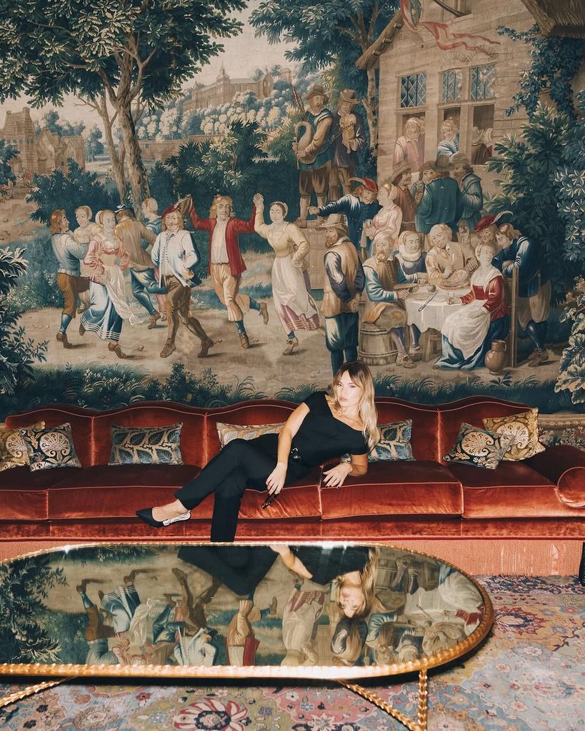 Lily James lounges on a deep red velvet couch in an all-black Dior ensemble, surrounded by an ornate 18th-century-style tapestry depicting a countryside celebration. The scene evokes old-world charm blended with contemporary fashion attitude.