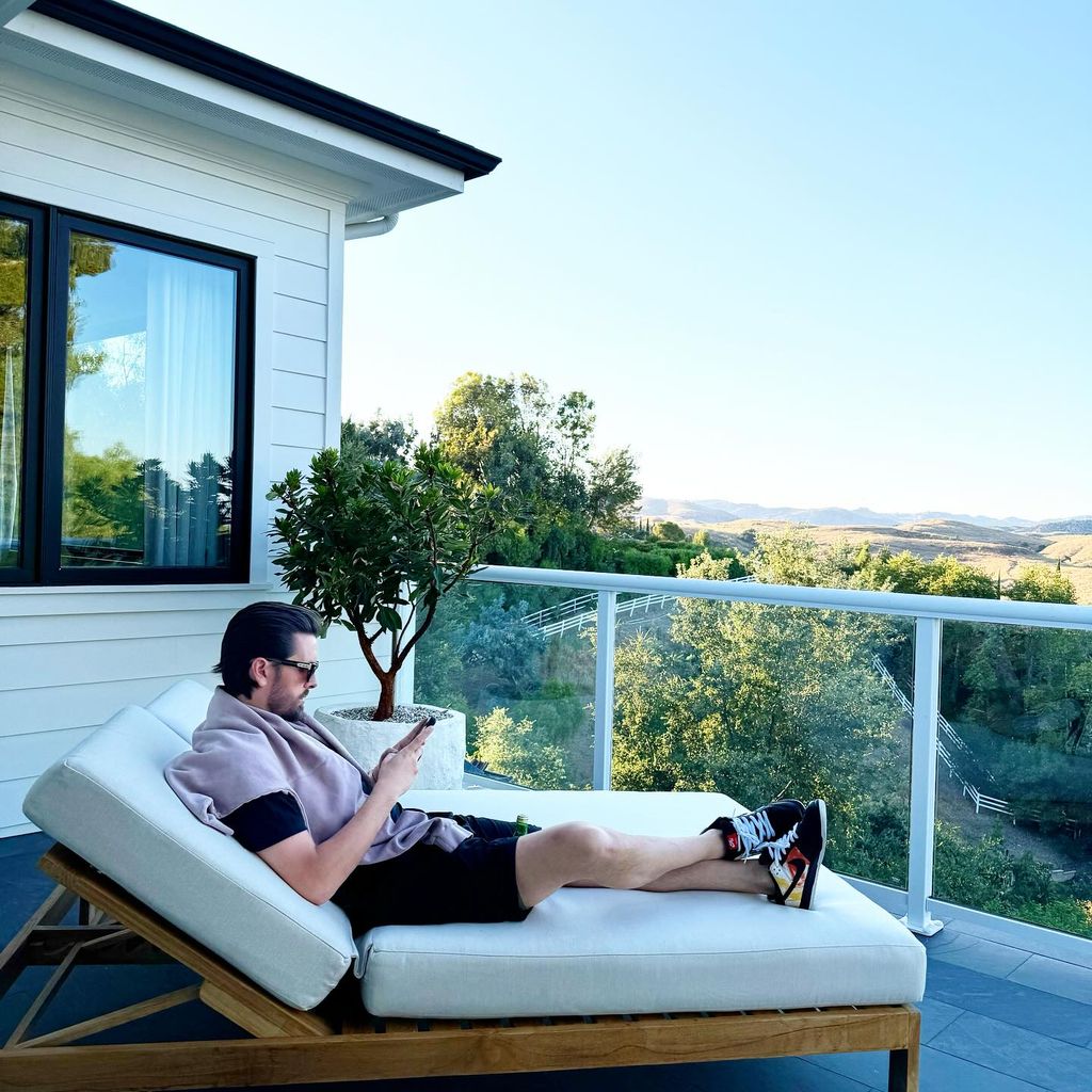 Scott Disick scrolling on his phone on a white sofa outside