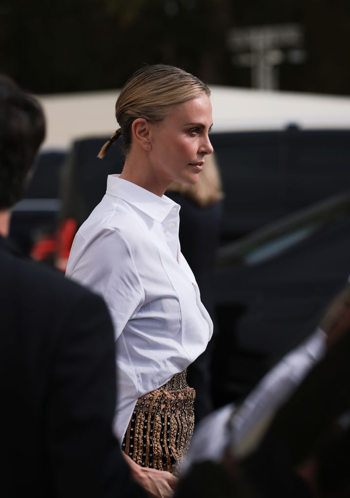 Charlize Theron is seen wearing a complete outfit from Christian Dior, consisting of golden logo ear studs with pearls; a white, buttoned, long-sleeved shirt; a see-through, high-waisted, brown, braided maxi skirt