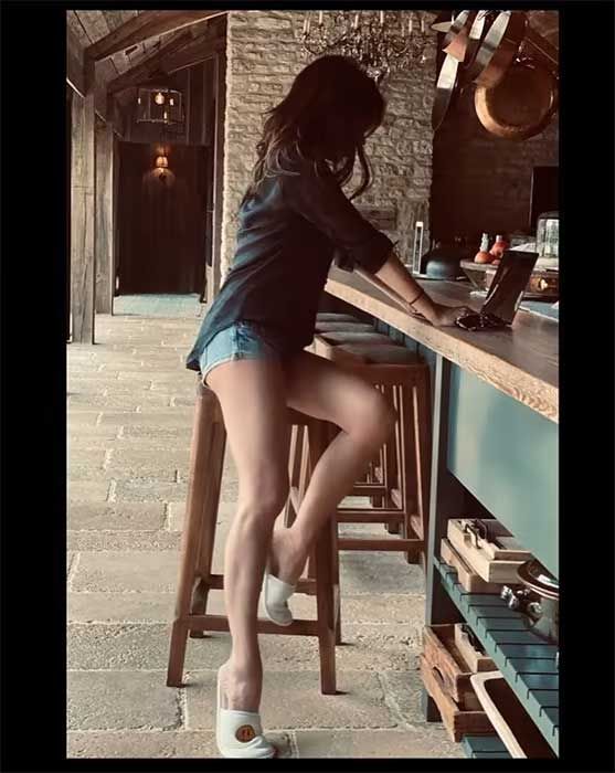 victoria wears hotpants and partially sits on a high stool in a huge openplan kitchen with bare stone work