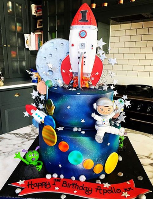 a two tier cake is decorated blue and black to look like the night sky and there are orange circles representing planets stuck to it with a little astronaut cartoon and rocket shaped cut out stuck to it 