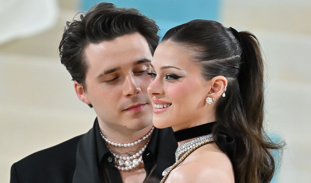 brooklyn beckham in a black suit with layered pearl necklaces gazes at nicola peltz wearing a black choker, diamond earrings, and dramatic winged eyeliner. The background is softly blurred with light blue and beige tones