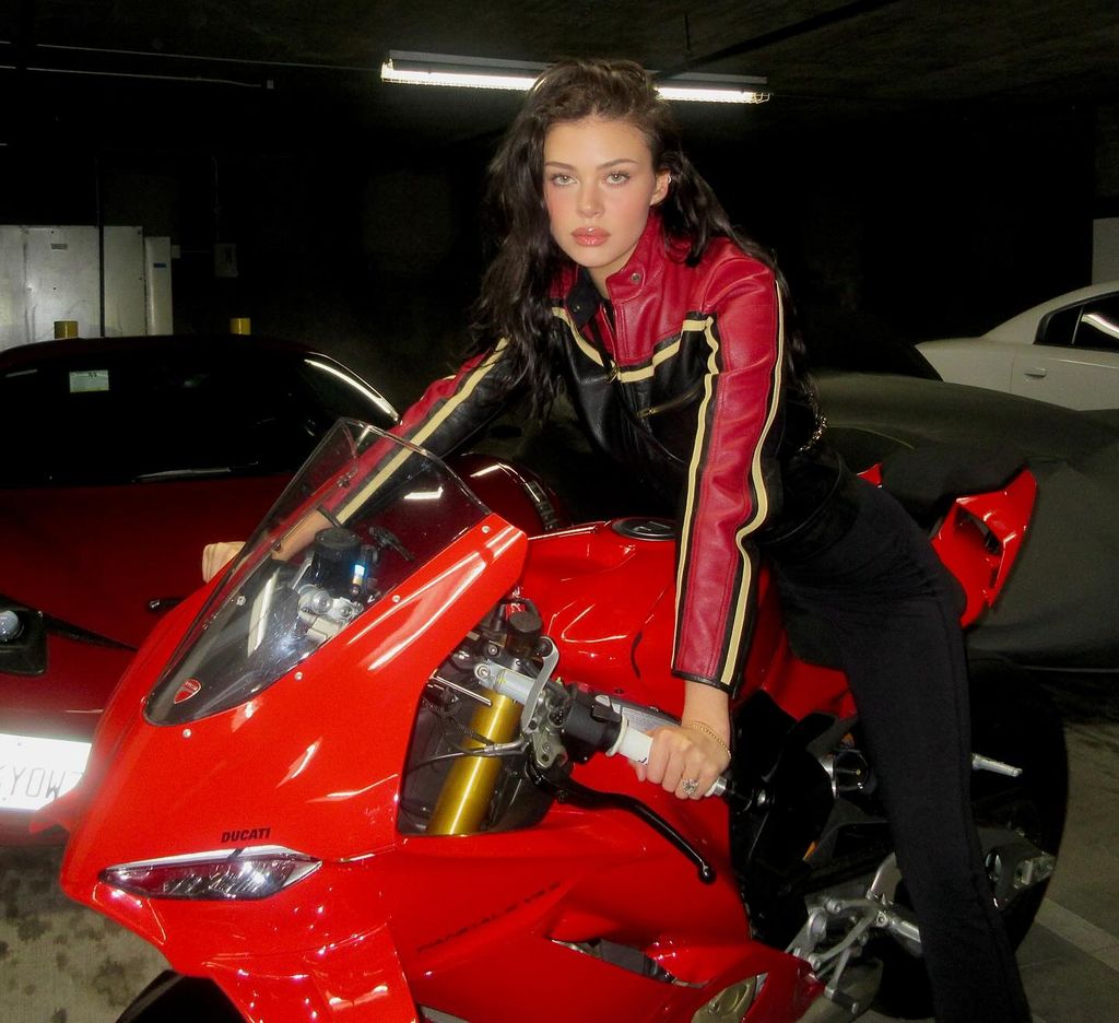 Nicola Peltz Beckham sits on top of a red motorbike, wearing a red, beige and black moto jacket and trousers