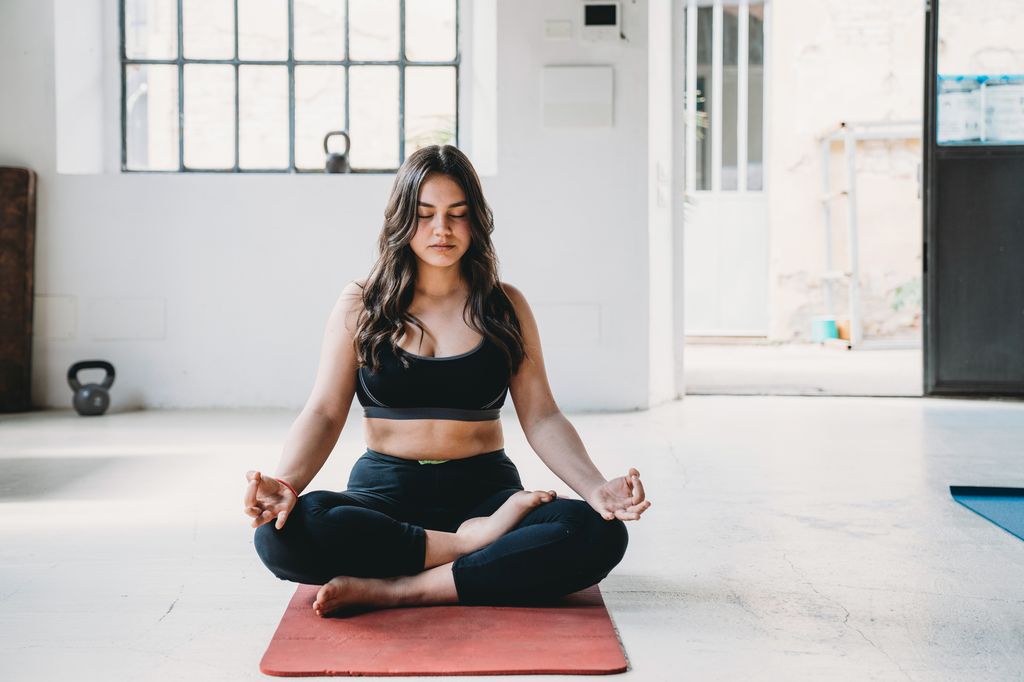Young adult woman is practicing yoga in a modern loft. Half lotus or ardha padmasana pose.