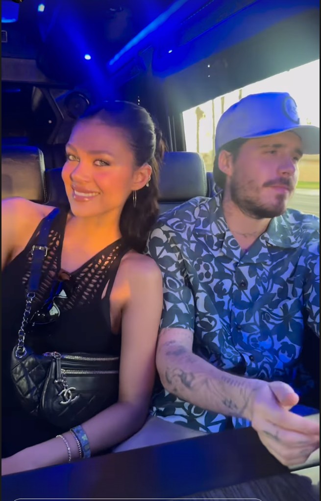 Nicola Peltz Beckham is seated beside Brooklyn Beckham wearing a blue patterned shirt and a light grey cap with the number "23" on it. Nicola is smiling radiantly, her long black hair swept back into a high ponytail, revealing hoop earrings and flawless makeup. Slung across her body is a black Chanel bumbag with signature quilted leather and silver zippers, its iconic charm swinging at her hip. Her black crochet-knit halter top reveals toned arms, adorned with delicate bracelets.