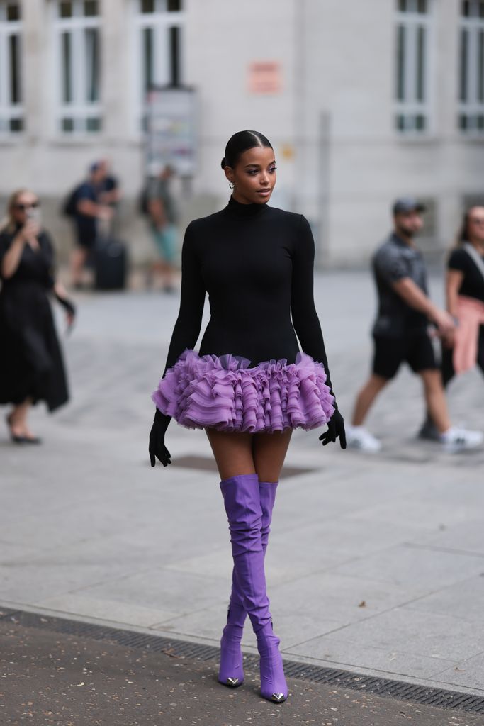 Ella Balinska is seen wearing silver hoop earrings, black sunglasses, a long-sleeved mini dress with black turtleneck and purple tulle tutu, black gloves and purple over knee boots with shiny silver toe caps during London Fashion Week September 2023 before the David Koma Show