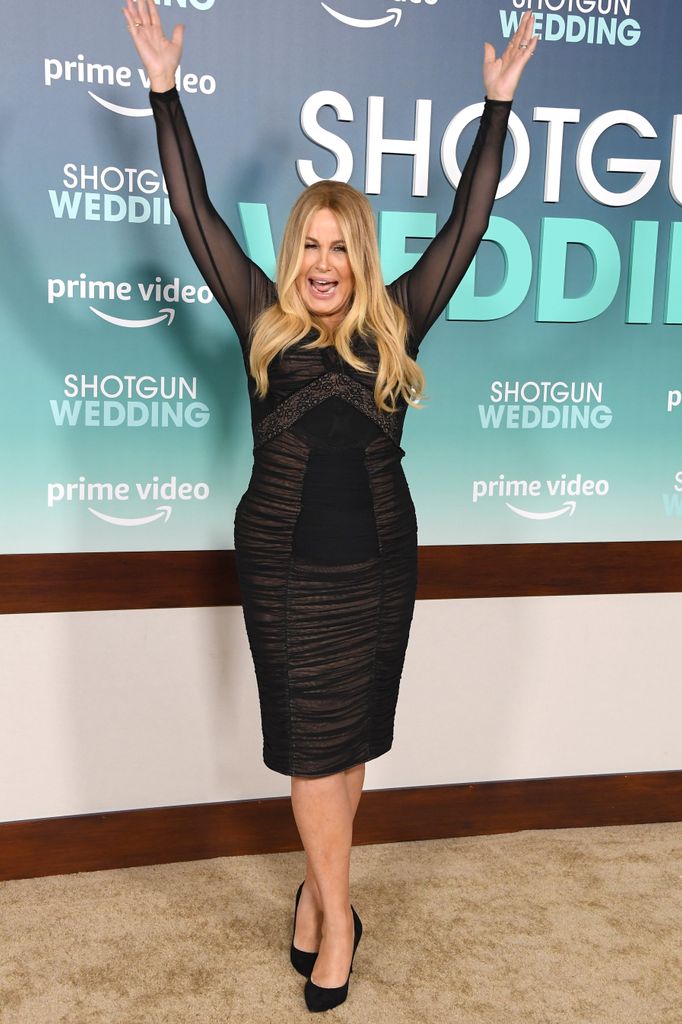 Jennifer Coolidge arrives at the Shotgun Wedding premiere. She is wearing a black knee length dress and high heels and is joyfully raising her hands above her head and smiling widely.