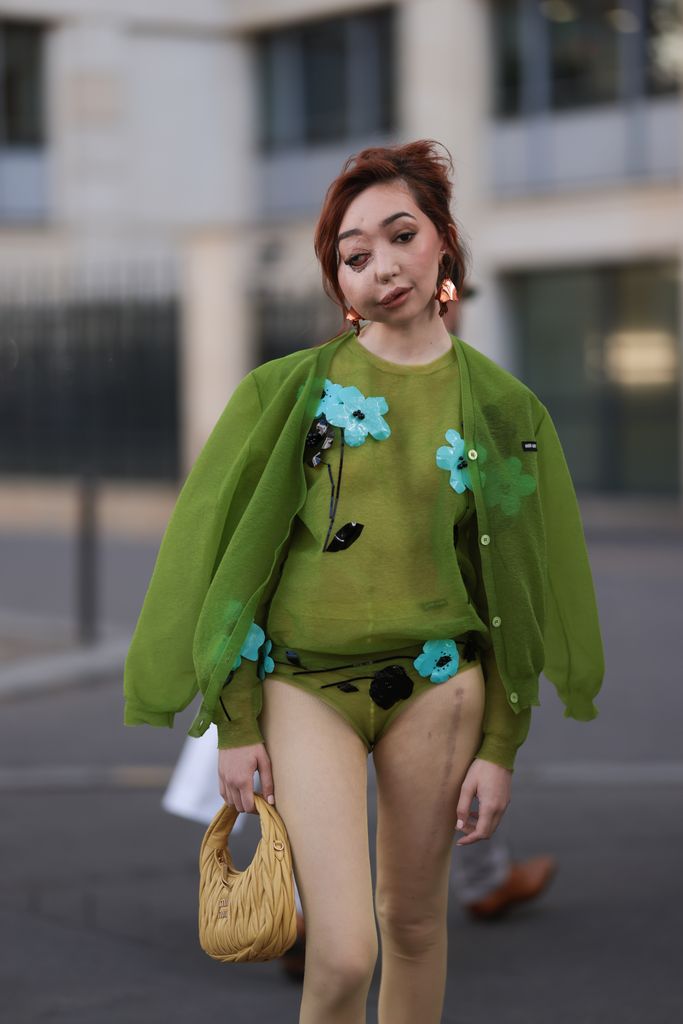 Nikki Lilly Christou is seen wearing a transparent green top with blue and black embroidered flowers, a green cardigan, a beige leather Hobo handbag by Miu Miu, white shorts by Miu Miu and a green shorts with embroidery, with black leather loafer during the Miu Miu Show Womenswear Spring/Summer 2024 