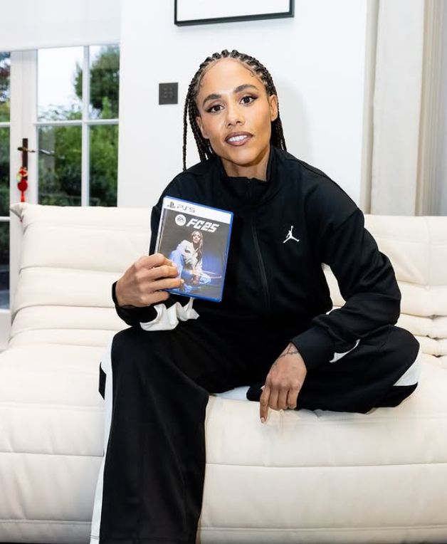 Alex Scott holding a copy of a football game while sitting on a cream sofa