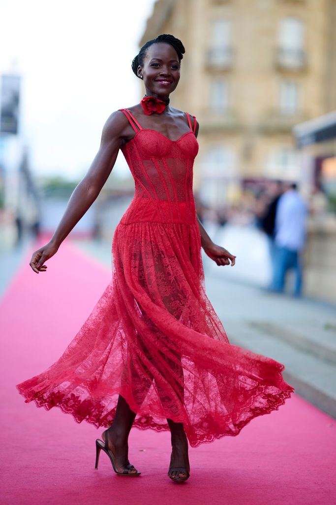 Actress Lupita Nyong'o woman twirls slightly, showcasing the movement of her red lace dress on the red carpet. The corset bodice and sheer flowing skirt catch the light. She smiles warmly, exuding confidence as the wind lightly plays with her dress.